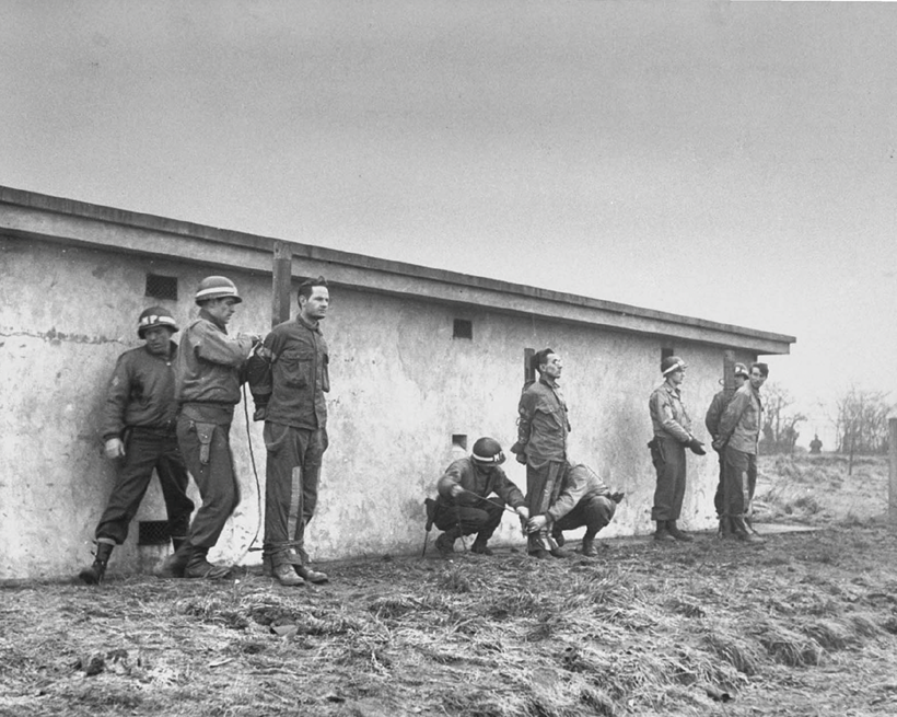 German infiltrators lined up for execution by firing squad after conviction by a military court for wearing U.S. uniforms during the Battle of the Bulge. December 23, 1944. 