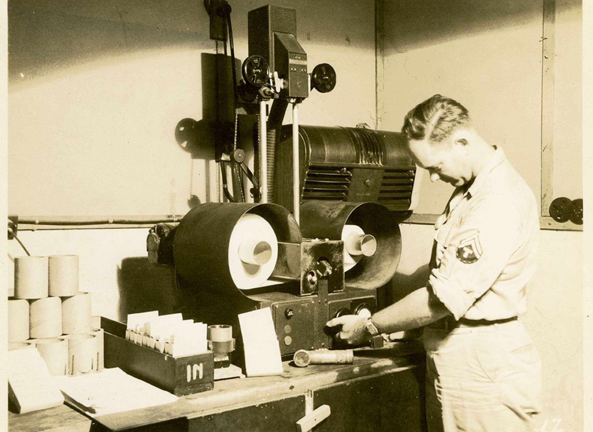 V-mail operation in the field at APO 929.”View of the continuous enlarger on which the image on the film is enlarged to regular V-Mail size of 4¼  x 5 inches. Sensitized rolls of paper, 825 feet in length are used. Here, the operator is adjusting the voltage to secure proper exposure. This is determined by consulting a chart which suggests certain voltage and lens aperture settings for any given film density.”