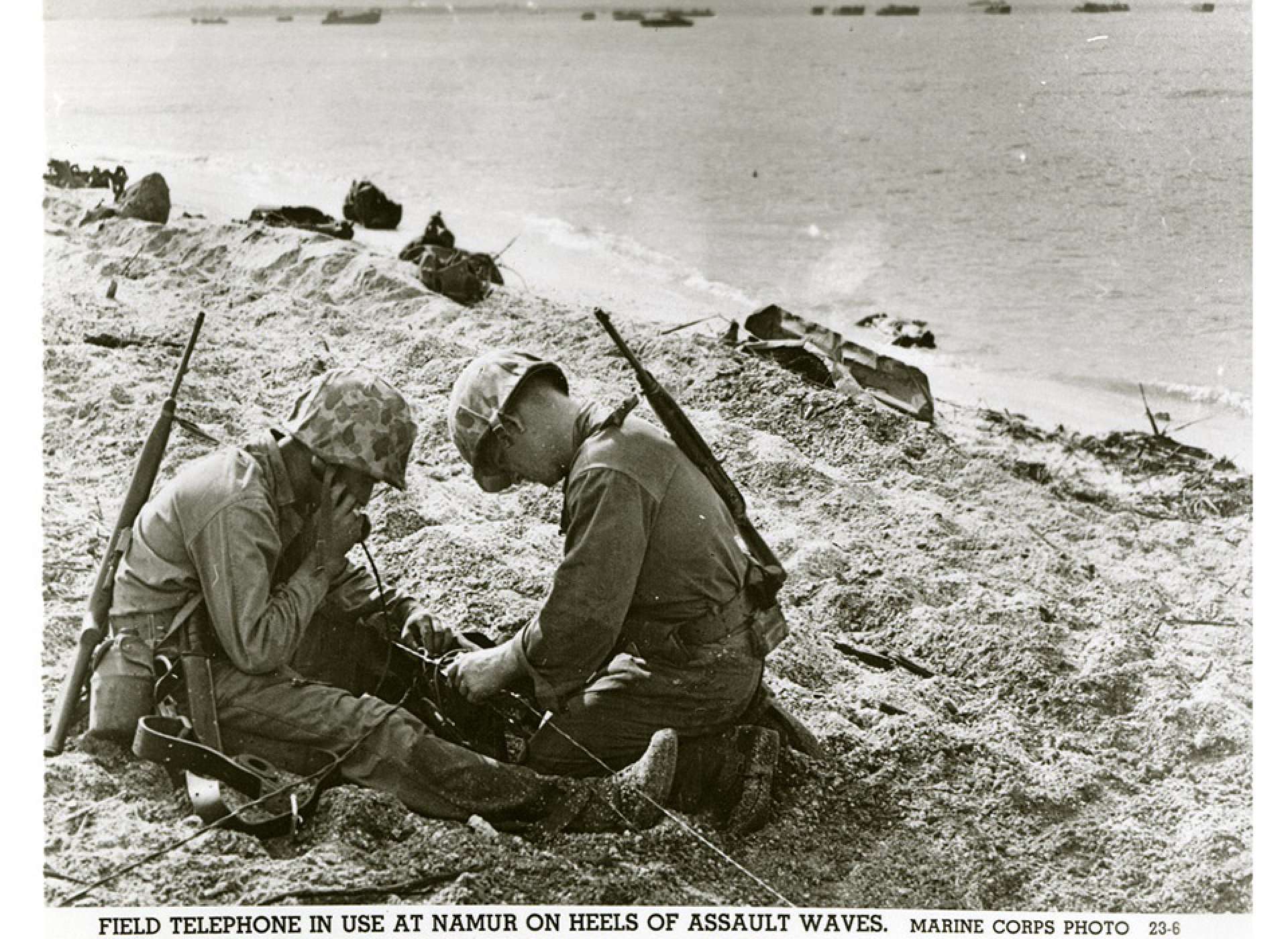 Two United States Marines using a field telephone on a beach; debris is scattered throughout; landing craft are in the ocean in the background. Official caption on front: &quot;Field telephone in use at Namur on heels of assault waves. Marine Corps Photo 23-6.&quot; Roi-Namur, Kwajalein Atoll, Marshall Islands. February 1944. Gift in memory of Sgt. Lyle E. Eberspecher, 2013.495.286