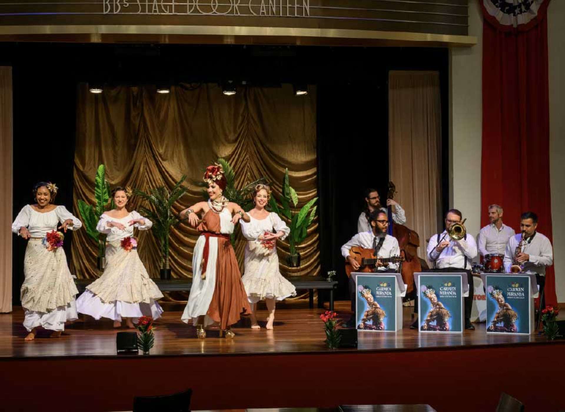 Four female dancers pose on stage in flowing white off-shoulder dresses and floral headdresses, with the lead performer in a rust-colored skirt and ornate jewelry striking a central pose against a gold draped curtain backdrop.