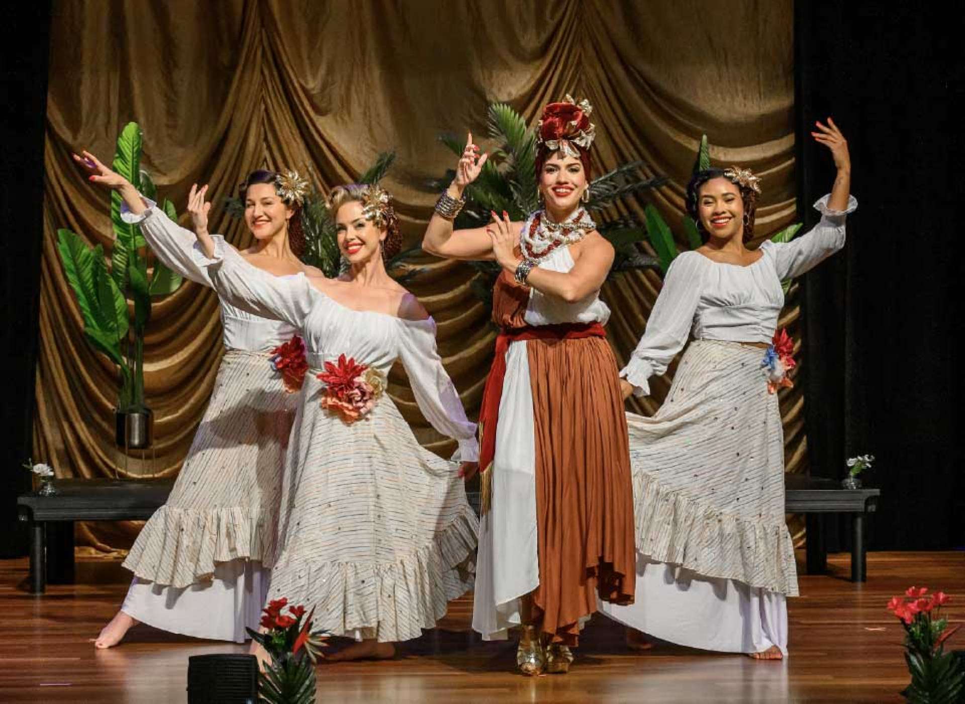 Four female dancers mid-performance on stage, with the lead in a rust and white dress and elaborate red floral turban headpiece extending her arms gracefully, while three supporting dancers in white ruffled skirts move around her against a gold curtain backdrop.